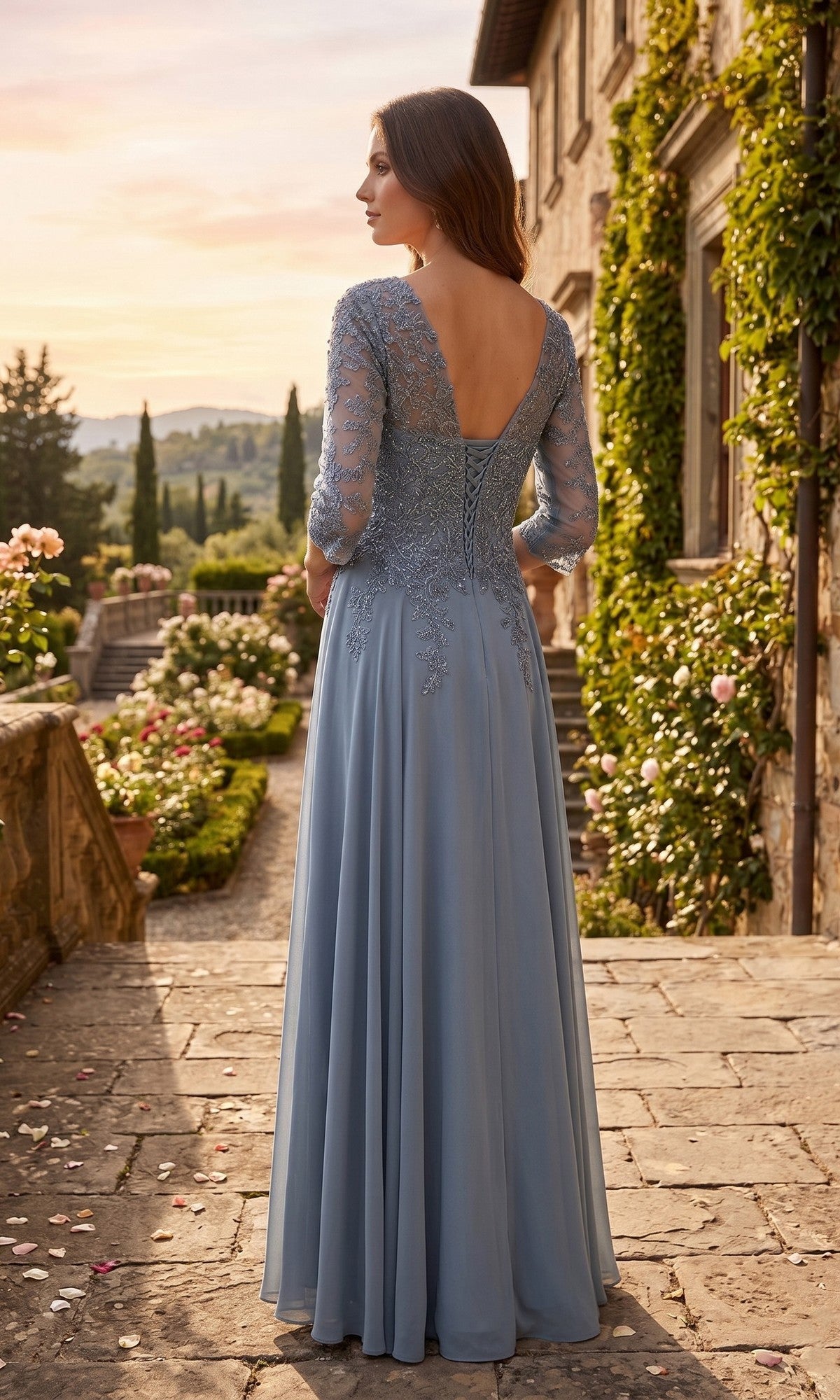 A woman wearing the Long Prom Dress: Chicas J070, a light blue chiffon evening gown with lace sleeves, stands outdoors on a stone path beside a greenery-clad building, facing away from the camera.